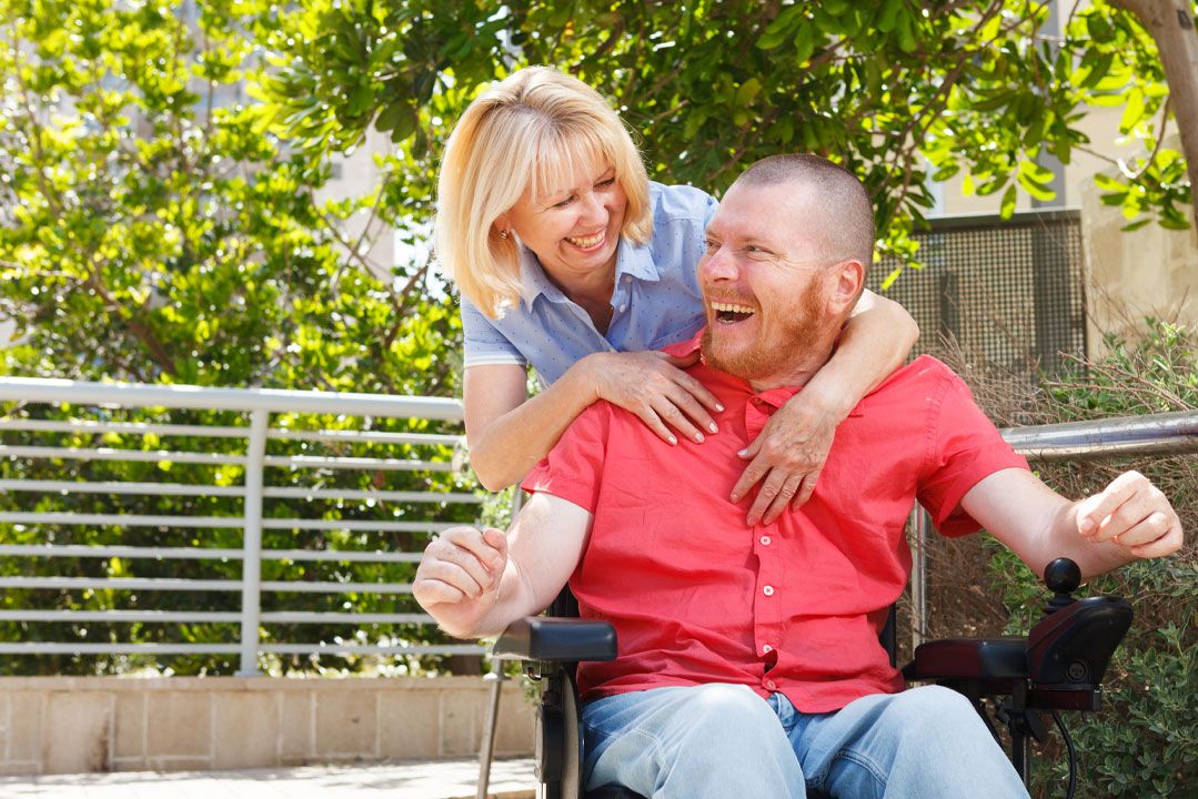 A man who uses a wheelchair laughing outdoors with a woman hugging him